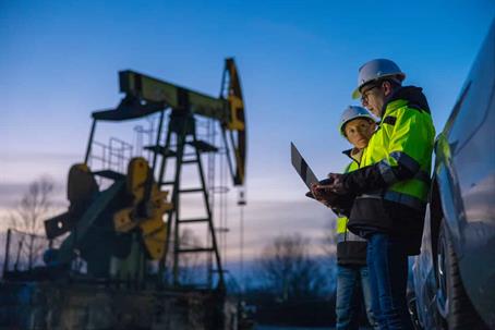 Two oilfield workers in high-visibility jackets are looking at a laptop while standing next to an inactive oil rig.
